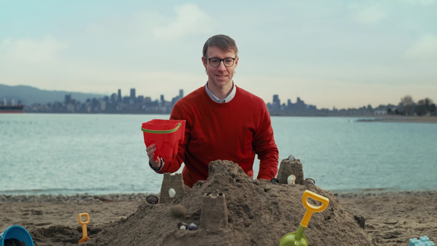 A man in a red sweater building a sandcastle with downtown Vancouver behind him, from Recouver in Vancouver